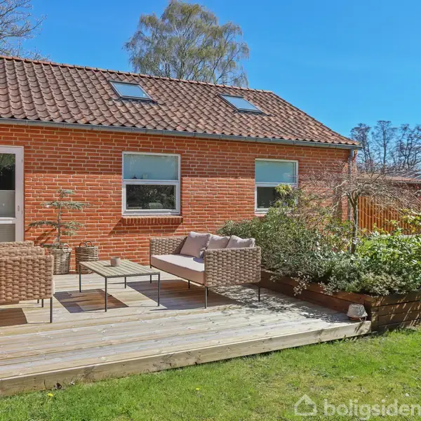 Rød murstenshus med rødt tag, terrasse med havemøbler og planter i træbeholder, omgivet af grøn græsplæne og træer i baggrunden.