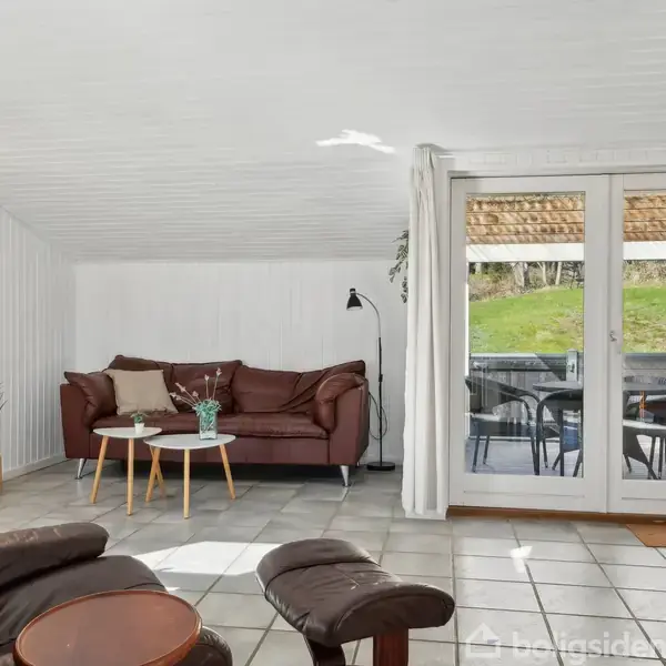 Stue med en brun lædersofa og rundt sofabord på klinkegulv. En lænestol med skammel står ved siden af. Store glasdøre åbner ud til en terrasse med udsigt over græsplæne. En gulvlampe og en malet kunstplakat i en gul vase afslutter indretningen.