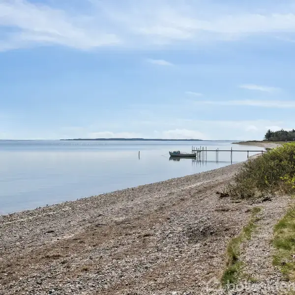 Kystlinje med småsten danner en bue langs stille hav, med en mole og båd. Grønne buske vokser tæt på vandet under en blå himmel.