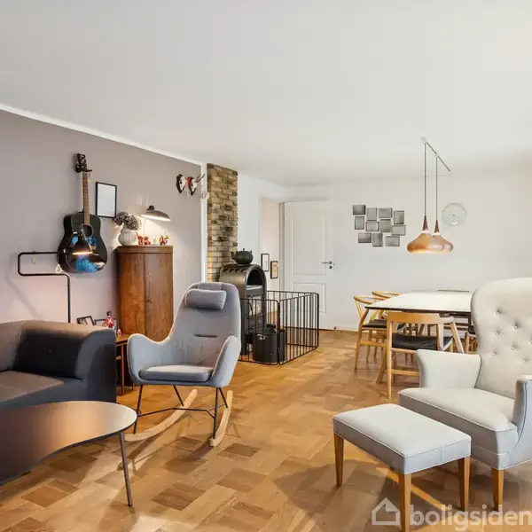 En stue med en sort sofa og en grå stol arrangeret omkring et bord. En guitar hænger på væggen. I baggrunden er et spisebord med stole og en brændeovn.