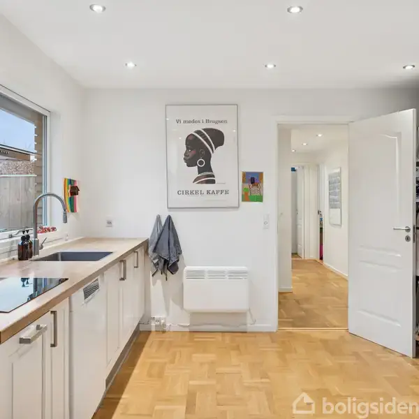 Køkken med indbygget ovn og kogeplade på en hvid bordplade. En plakat med teksten "Vi ønsker i Begynde CIRKEL KAFFE" hænger på væggen. Parketgulv og åbent dørområde.