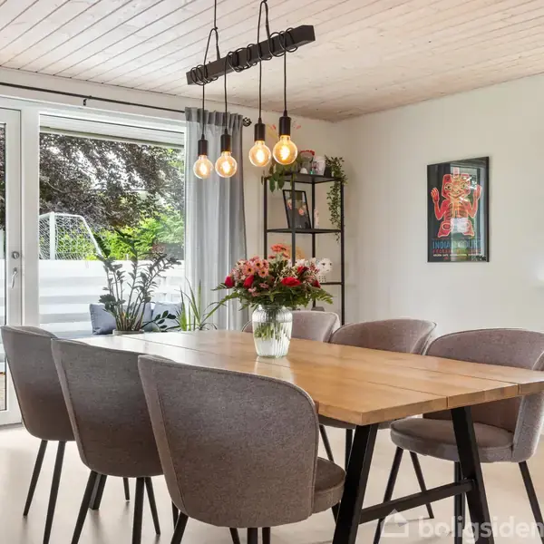 Et spisebord med fire stole står i et moderne, lyst spisestueområde. En vase med blomster er placeret på bordet. Store vinduer og en glasdør fører ud til en terrasse med planter.