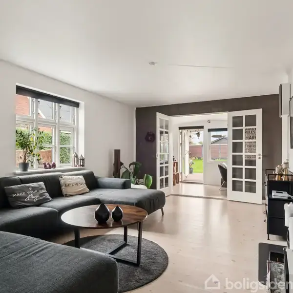 Stuen har en stor, grå sofa med puder, placeret foran et rundt sofabord. Dobbelt franske døre åbner ud til en lys gang. En pejs er til højre.