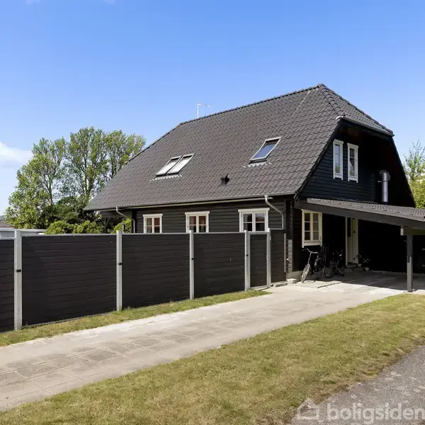 Sort hus med skråtag står på en grund omringet af græs og hegn. Der er en indkørsel ved siden af huset og et overdækket område. Himlen er blå og klar.
