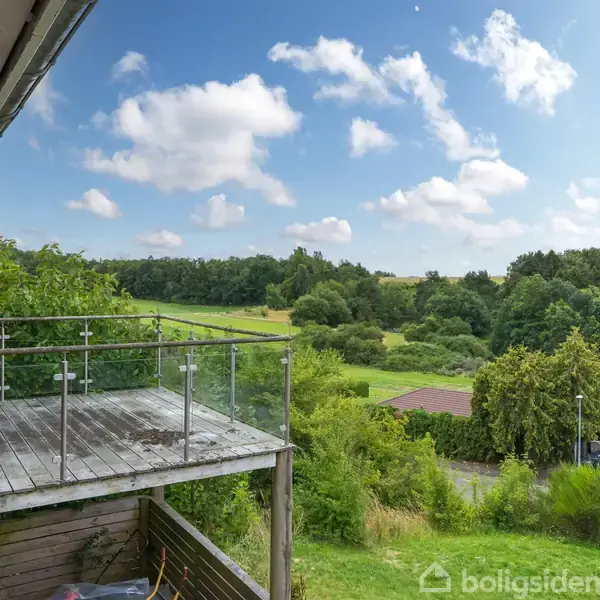 En træterrasse med glasrækværk hænger ud fra et hus, mod en frodig grøn udsigt med skov og marker under en skyet blå himmel.