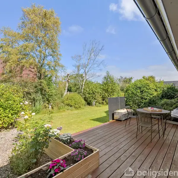 Terrassen er hovedfokus og indeholder et bord med stole. Den er placeret ved et murstenshus med udgang til en grøn have fyldt med buske og et træ under en blå himmel.