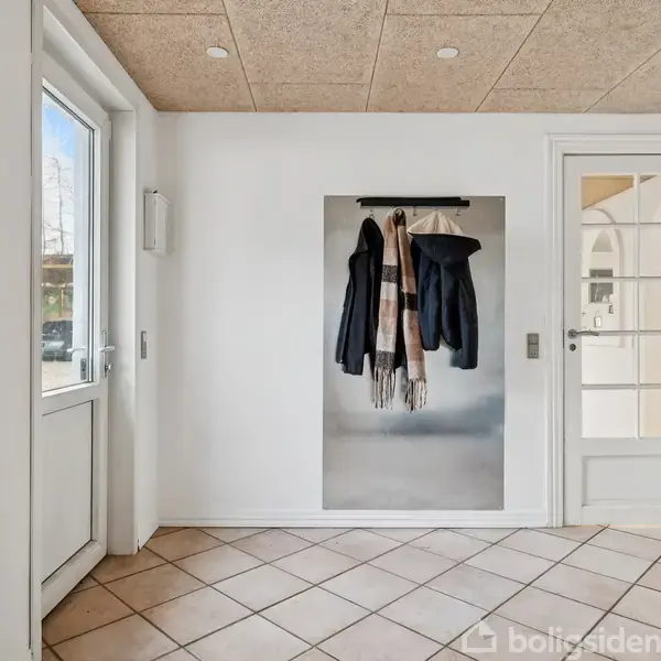 Dør med jakker og tørklæde hængende på en knage på en hvid væg. Flisegulv og glasdør i baggrunden. Lys indtrænger gennem vindue til venstre.