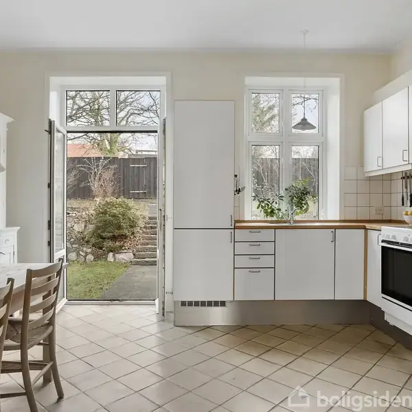 Badeværelse med et hvidt, flisebelagt badekar ved et vindue. En hvid skabsvæg står til venstre, og et håndklæde hænger over en vaskemaskine. Vinduet viser et træ udenfor.