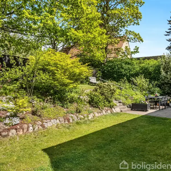 Et lyst rum med trægulv rummer en bogreol fyldt med bøger, mens et rundt spisebord er omgivet af stole. Store vinduer lukker naturligt lys ind.
