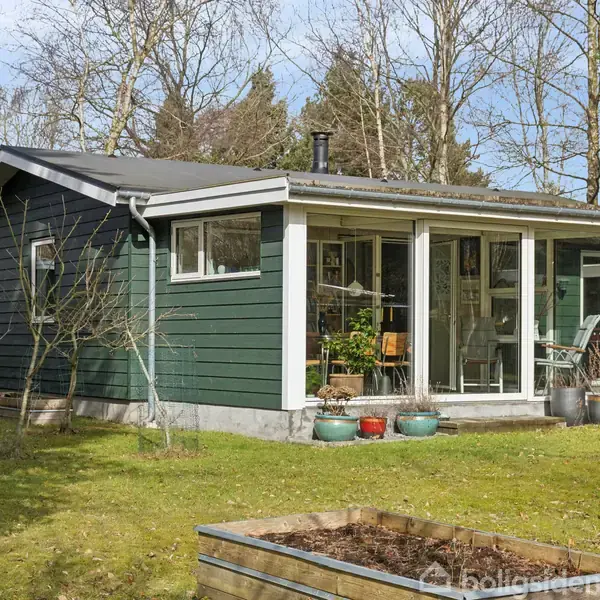 Et lille, grønmalet sommerhus står på en græsplæne omkranset af træer. Huset har en veranda med glaspartier, potteplanter foran, og en planteramme på græsset.