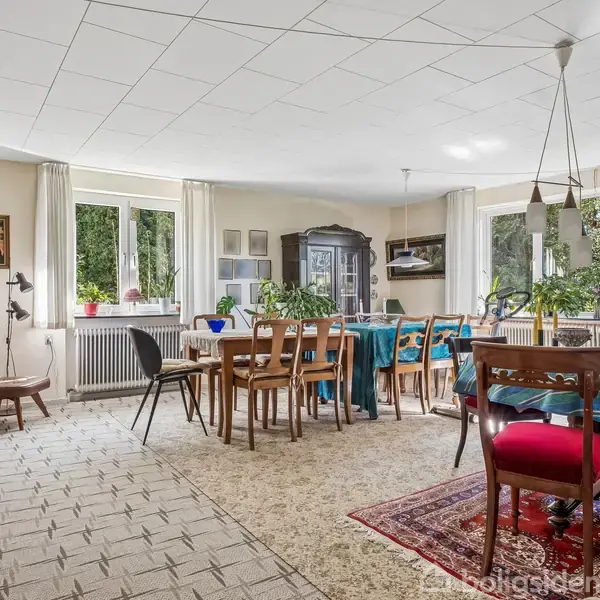 Spisestue med et langt træbord omgivet af stole. Bordet er dækket med en blå dug. Planter på bordet og i vindueskarmen. Værelset har store vinduer og tæppebelagt gulv.