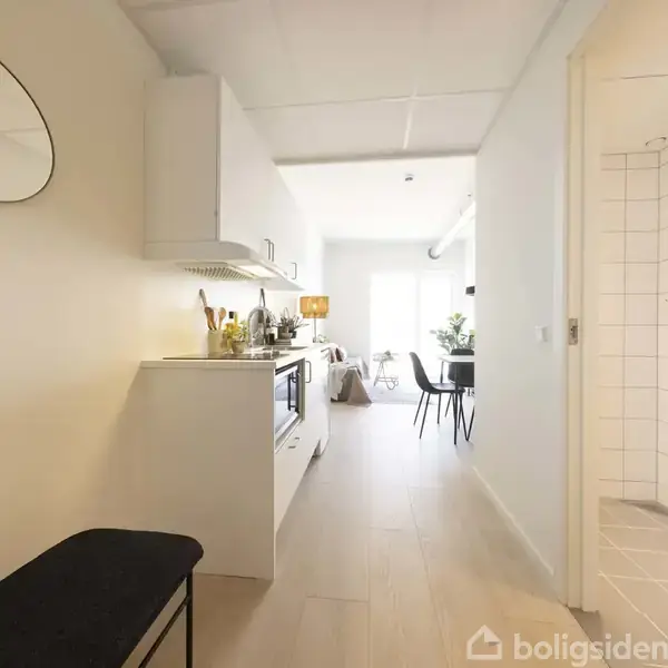 Køkken-alrum med hvidt interiør fører til en spisestue med stole og et bord. Øverst til højre ses en indgang til en enkel bruser med hvidt flisebelagt væg.