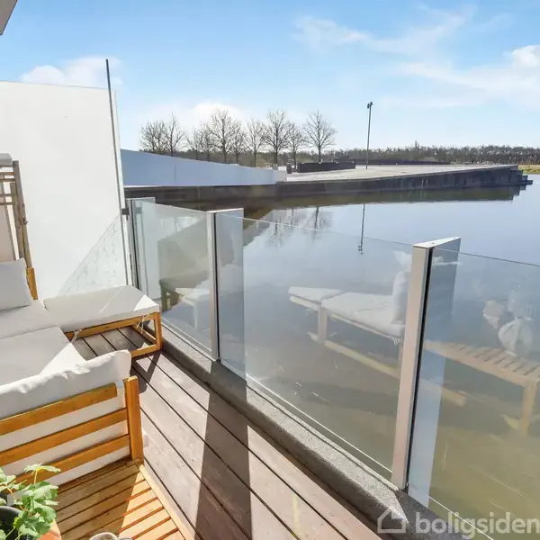 En balkon med en hvid sofa og et lille bord står ved glasrækværk, spejlende i en rolig sø. Omkringliggende træer skaber en fredfyldt atmosfære på en solrig dag.