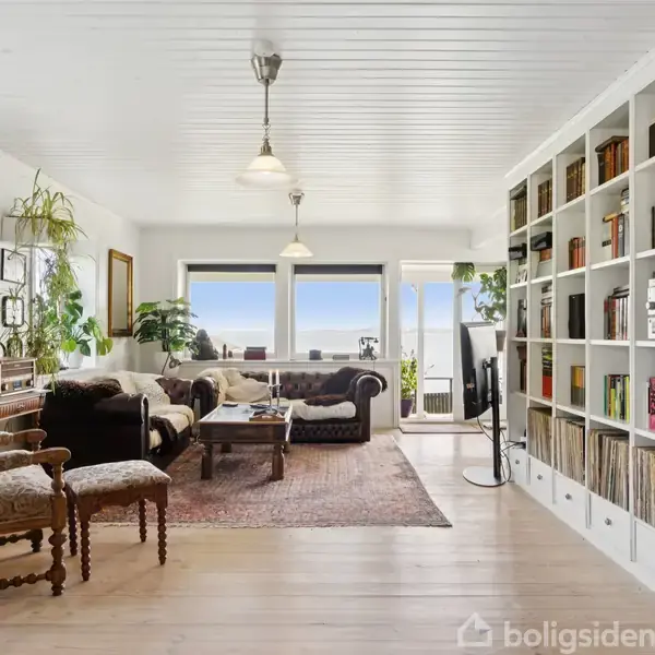En stue med sofaer og et sofabord står foran vinduer med udsigt til havet. Bøger fylder en stor reol til højre. Planter og dekorative billeder pryder væggene.