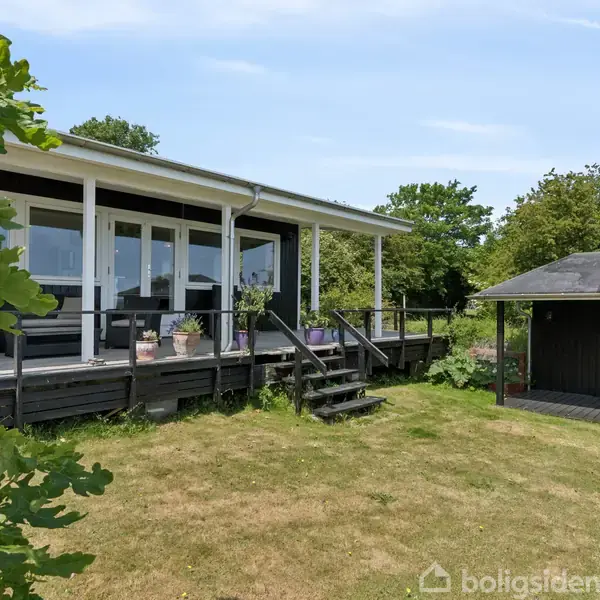 Sommerhus med stor terrasse står stille i en grøn have med træer og buske omkring. En lille, sort udbygning ligger til højre nær græsplænen.