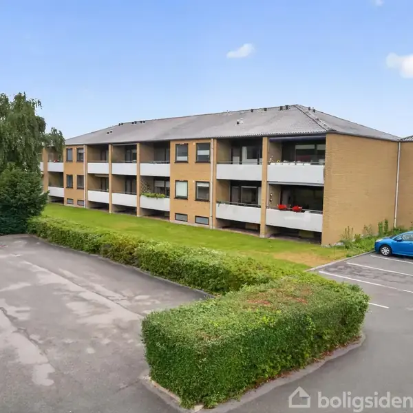 En gul murstensbygning med altaner står stille ved siden af en parkeringsplads, der har en parkeret blå bil og grønne buske i nærheden under en let skyet himmel.