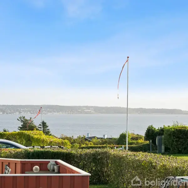 Flagstang med hvidt flag vajer i brisen ved siden af en have indrammet af hække. Udsigt over en bugt med fjerne kystlinjer under en klar blå himmel.