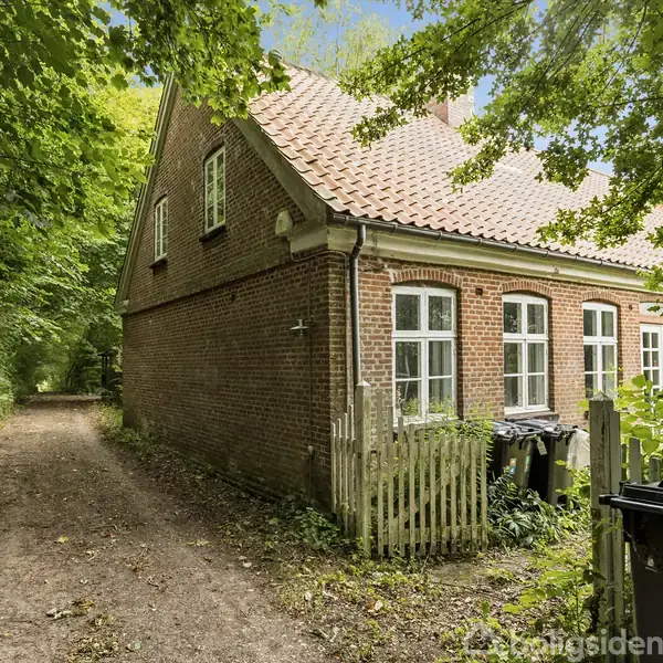 En murstensbygning med rødt tag og hvide vinduer står ved en grussti omgivet af grønne træer. Foran huset er der skraldespande og et lille træhegn.