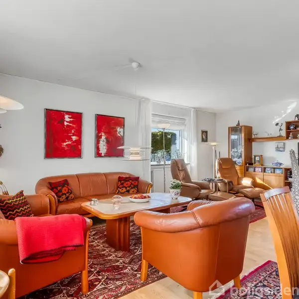 Spisebord med stole står på et rødt tæppe i stue-alrum. Stuen har et brunt hjørnesofa-arrangement. Store vinduer med udsigt til have. To røde billeder hænger på væggen.