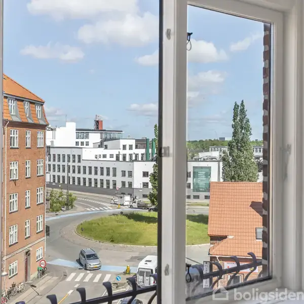 Et åbent vindue med udsigt over en rundkørsel. Der er røde og hvide bygninger i baggrunden, samt blå himmel med skyer.