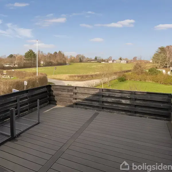 En stor, sort terrasse med moderne rækværk står stille i forgrunden, omgivet af åbne grønne marker og spredte træer under en blå himmel med hvide skyer.