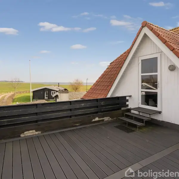 En hævet terrasse med trin foran et hvidt hus med rødt tagår. Terrassen har sort rækværk og trægulv. I baggrunden ses en åben mark og en skyfri blå himmel.
