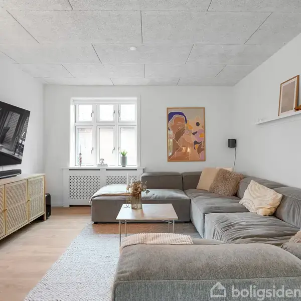 Stor grå sofa står i en lys stue med et stort vægmonteret fjernsyn og en abstrakt plakat. Et sofabord står foran sofaen, og et lille vindue lader lys ind.