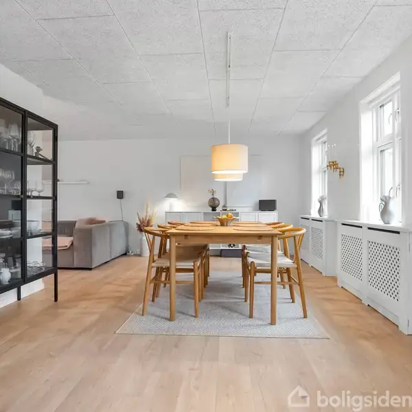 Spisebord med seks stole står midt i en lys stue. Ved siden af er der et vitrineskab, og et stort vindue lyser rummet op. En sofa står i baggrunden.