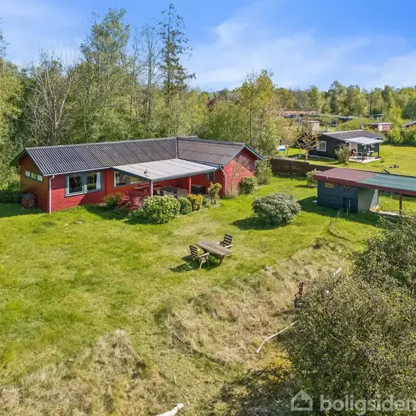 Rødt træhus står stille i en grøn have med græsplæne og borde. Omgivet af træer og åbne områder, ligger huset i et landligt, skovfyldt område.