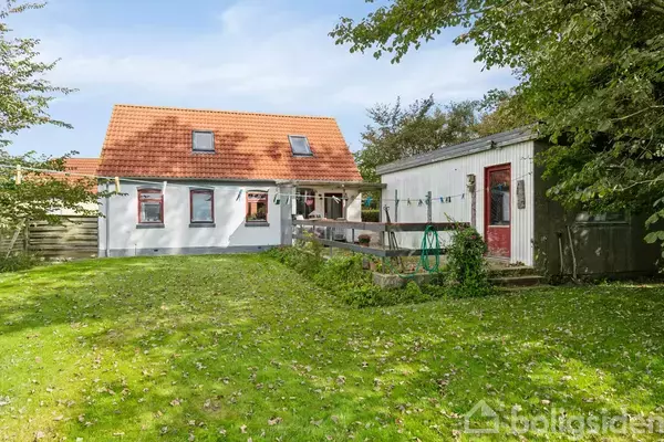 Hus med rødt tegltag har hvid facade og flere små vinduer. I haven er der en græsplæne og en skurbygning med en rød dør. Træer omkranser området.