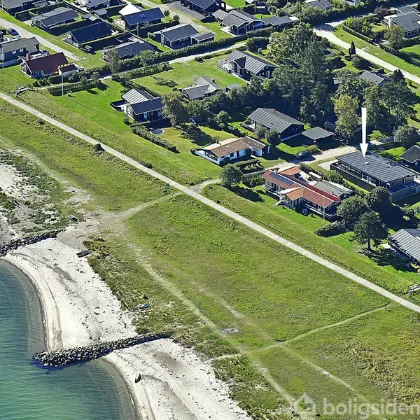 Kystnære huse ligger i en række nær en grøn sti med en strand til venstre. Husene varierer i størrelse og er omgivet af træer og haver.