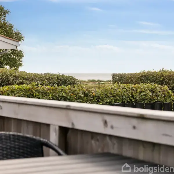 Et udendørs træbord med en kurvestol står på en terrasse. Baggrunden viser buske og huse med synlig himmel og hav.