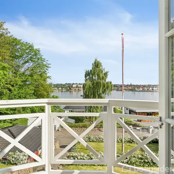 Åben altandør giver udsigt over en sø, med træer og et hus tæt ved. Det er solrigt med klare blå himmel.