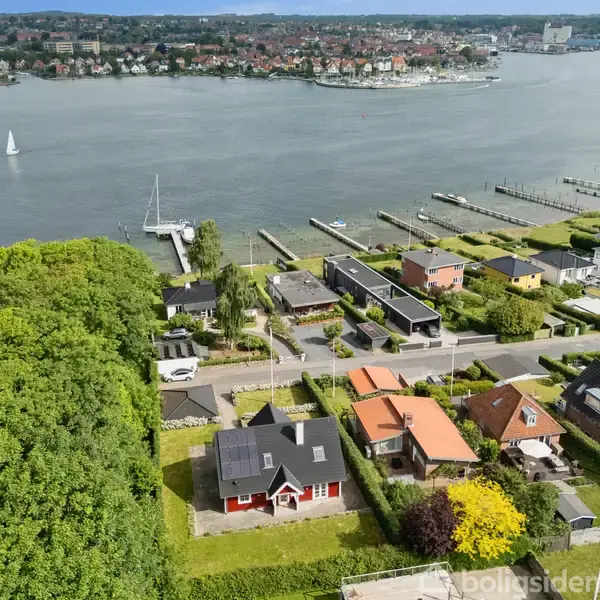 Et hus med solpaneler står i en grøn have. Det ligger tæt ved en vej, med flere huse og en marina i nærheden. En bred flod ses i baggrunden.