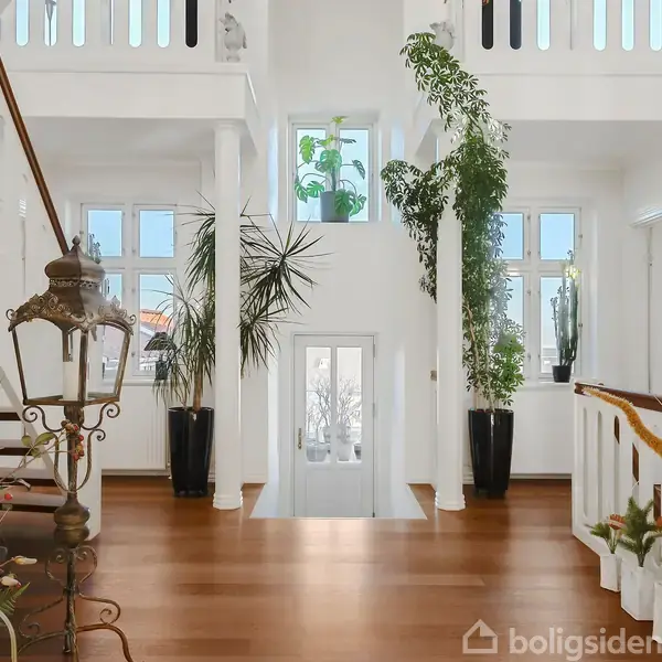 Et lyst rum med trappe og planter, der står i krukker. Trappen har hvidt gelænder, og rummet har lyse vægge og vinduer, der slipper masser af lys ind.