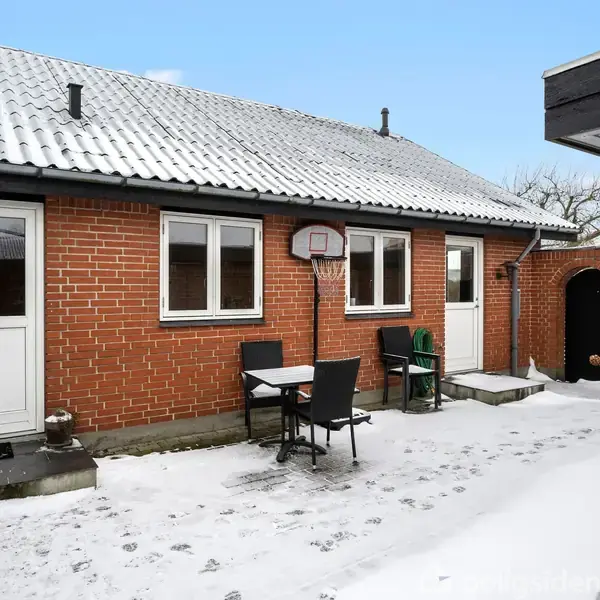 En rødstensbygning med fladt tag har to hvide døre og flere vinduer. En basketballkurv hænger på væggen. En gårdhave med sne og et bord med stole ses foran.