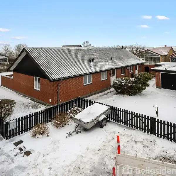 Rødt murstenshus med sort tag, omgivet af vinterlandskab med sne. En trailer står stille ved et sort stakit. Til venstre ses flere huse i baggrunden.