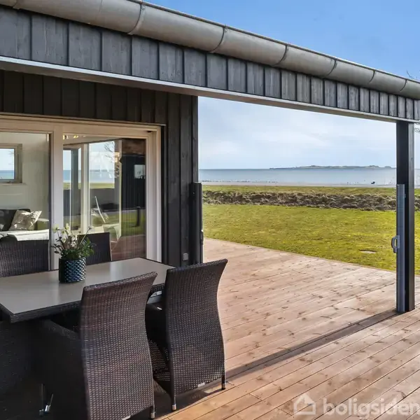 Terrassebord med flettede stole står på en træterrasse ved et sommerhus. I baggrunden ses en græsplæne, træer og en udsigt over vandet under en blå himmel.