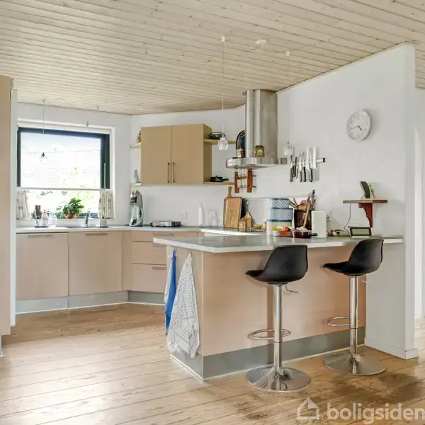 To moderne barstole står ved en køkkenø, omgivet af et lyst køkken med åbne hylder, køkkenudstyr og et vindue. Væggene er hvide, og gulvet er i træ.
