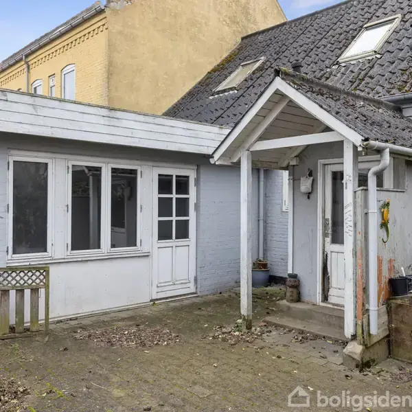 Et hvidt hus med gråt tag har en overdækket indgang. Der er vinduer ved siden af hoveddøren. I gårdspladsen står en lanterne og en trækasse ved siden af døren.