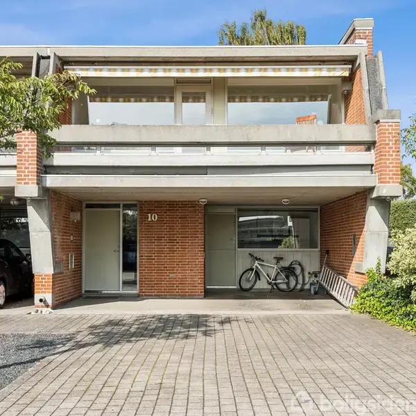 To-etagers rækkehus i mursten har en carport og en balkon. En bil og en cykel er parkeret foran. Huset har nummeret 10 og er omgivet af træer og buske.