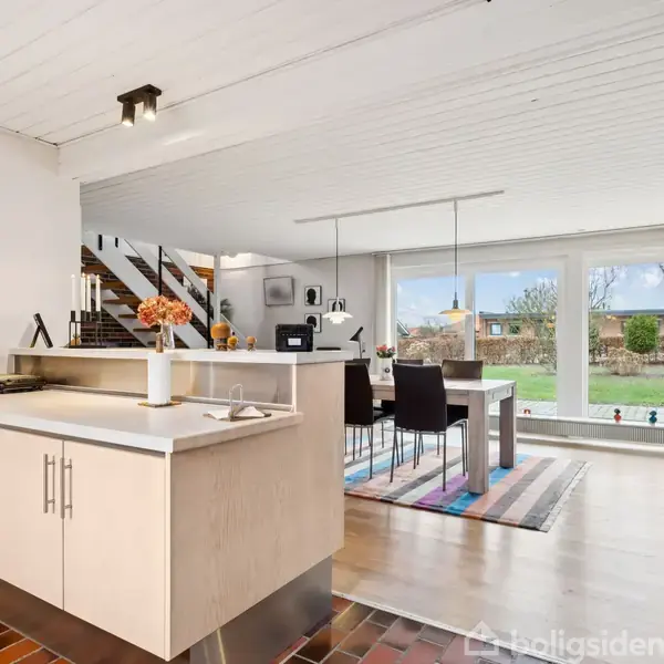 Køkkenbord med vase og lys, stående på mørkt trægulv. I baggrunden ses et spisebord med stole foran store vinduer, der afslører en have udenfor.