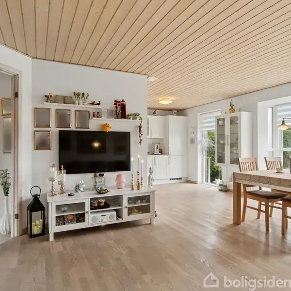 TV på en hvid reol viser et moderne rum. Spisestue ses i baggrunden med træbord, stole og store vinduer. Lys træindretning skaber en lys og åben atmosfære.