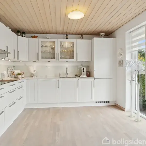 Et lyst, moderne køkken har hvide skabe og skuffer, integrerede apparater og dekorative elementer. Det er oplyst af indbygget lys og har store vinduer, der åbner mod en haveudsigt.