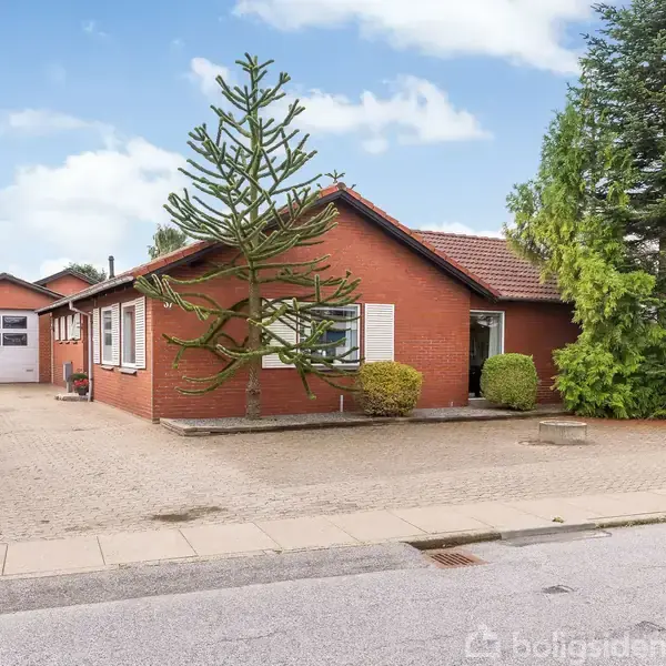 En rød murstensbungalow med fladt tag står omkranset af buske og træer. I forgrunden er en stor, flisebelagt indkørsel ved siden af huset.
