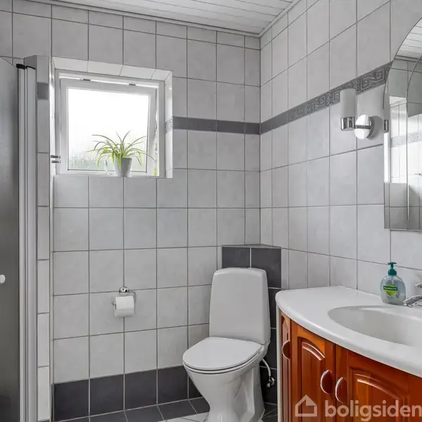 Toilet ved siden af en håndvask med sæbedispenser, omgivet af lyse fliser. Et lille vindue med en plante ovenpå står over toilettet, og en bruser ses til venstre.