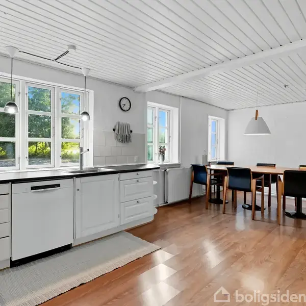 Køkkenet har hvide skabe og en sort bordplade. Et spisebord med stole står ved et vindue. Trægulve og hvidt træloft skaber en lys atmosfære. En ur hænger på væggen.