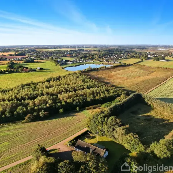Hus omgivet af frodige marker og træer, set fra oven. I baggrunden ses en bygd med en stor sø, under en klar blå himmel.
