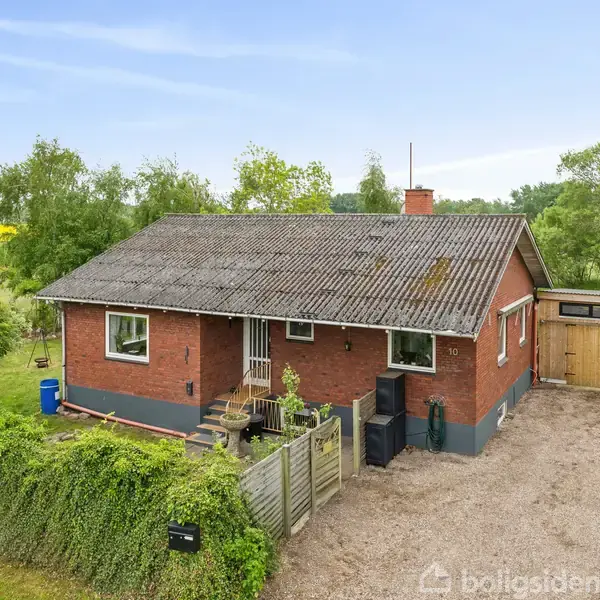 Rødt murstenshus med gråt tag står stille i en grøn, landlig have. Til højre er en grusbelagt indkørsel med carport. Husnummer "10" vises ved døren.