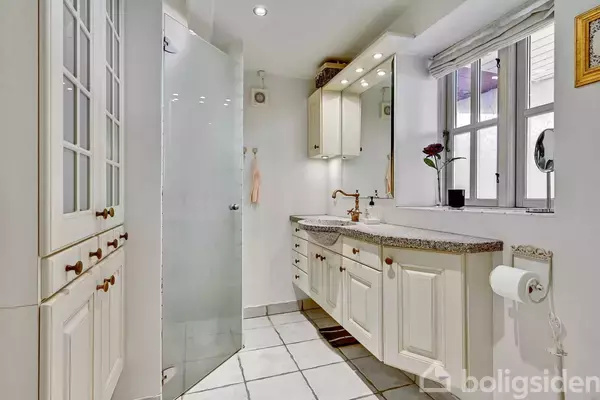 Badeværelse med hvidt skab og spejl over granitvasken, bruser med glasvæg til venstre. Vindue med blomster i karmen til højre. Flisegulv understreger det lyse rum.